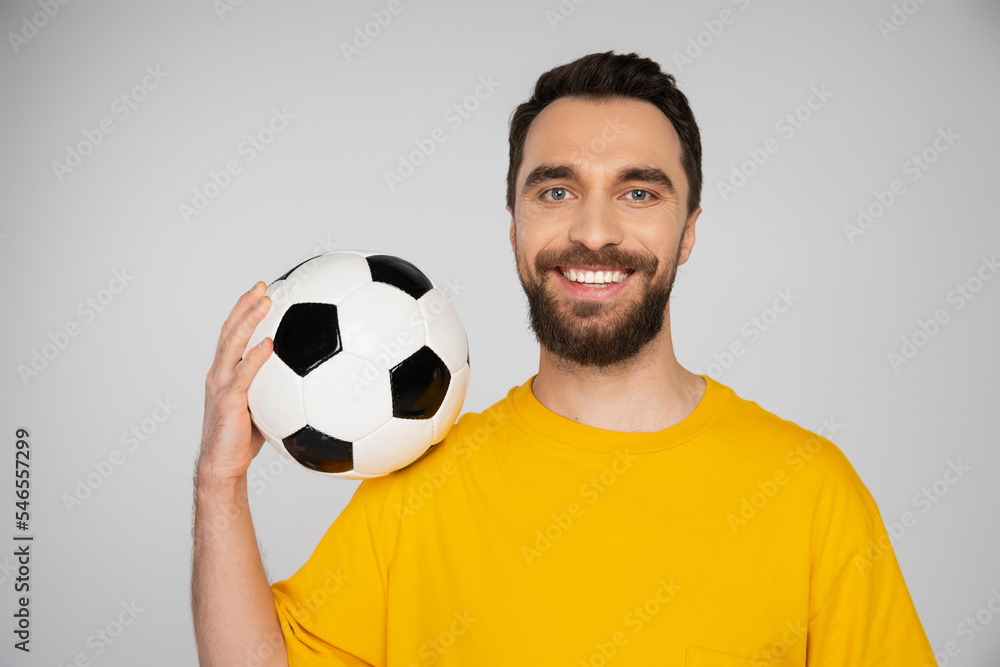 pleased bearded football fan in yellow t-shirt holding soccer ball and looking at camera isolated on grey. pleased bearded football fan in yellow t-shirt holding soccer ball and looking at camera isolated on grey.