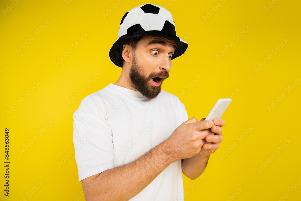 shocked man in football fan hat watching championship on cellphone isolated on yellow.
