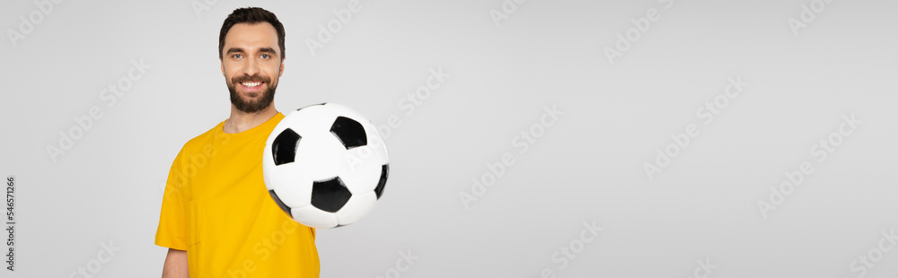 happy and bearded sports fan showing soccer ball and looking at camera isolated on grey, banner. happy and bearded sports fan showing soccer ball and looking at camera isolated on grey, banner.