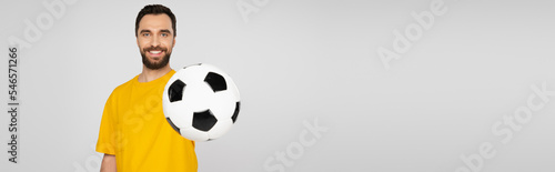 happy and bearded sports fan showing soccer ball and looking at camera isolated on grey, banner.