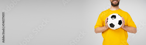 cropped view of bearded and smiling man in yellow t-shirt holding soccer ball isolated on grey, banner. © LIGHTFIELD STUDIOS
