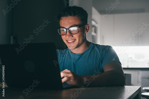 One young teenager studying and working at home at night on the table using laptop pc smiling and having fun doing homework. Light of screen on face..