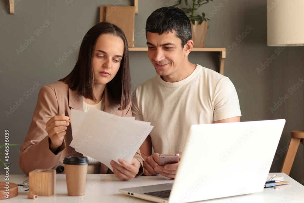Portrait of smiling man and serious concentrated woman sitting at table together in front of laptop working together, female holding papers, analyzing data. Portrait of smiling man and serious concentrated woman sitting at table together in front of laptop working together, female holding papers, analyzing data.