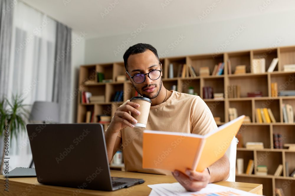 Arab man studying online from home, reading notebook and drinking coffee, sitting at desk with laptop pc, free space Arab man studying online from home, reading notebook and drinking coffee, sitting at desk with laptop pc, free space