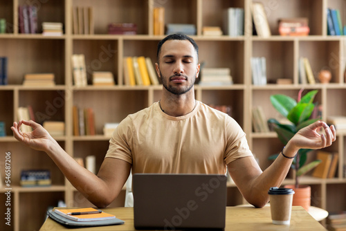 Workplace stress management. Calm arab man meditating with closed eyes in front of laptop, sitting at desk at home photo
