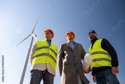 Wind turbines operate behind specialists of maintenance. Senior engineer and technicians look around working wind farm low angle shot