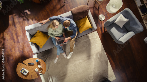 Top View of Loft Apartment: Happy Couple Using Laptop in the Stylish Living Room. Looking at Digital Display. On Couch: Boyfriend Hugs Girlfriend, They Use Computer for Online Shopping, e-business.