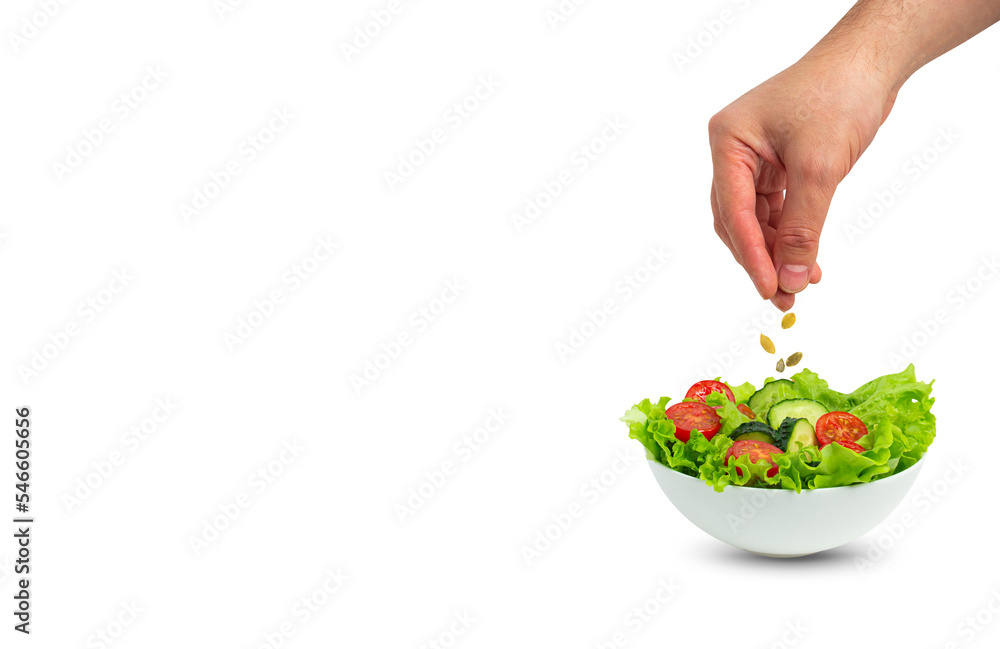 White bowl of salad with fresh cucumbers, tomatoes, iceberg salad leaves and male hand sprinkling pumpkin seeds over salad isolated on green background, template for advertising, space for text
