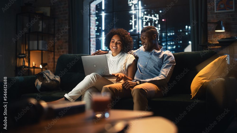 Multiracial Couple in Love Use Laptop Computer, while Sitting on the Sofa in Stylish Loft Apartment. Boyfriend and Girlfriend Buy Goods in Internet, Using Social Media Services, Chatting with Friends. Multiracial Couple in Love Use Laptop Computer, while Sitting on the Sofa in Stylish Loft Apartment. Boyfriend and Girlfriend Buy Goods in Internet, Using Social Media Services, Chatting with Friends.