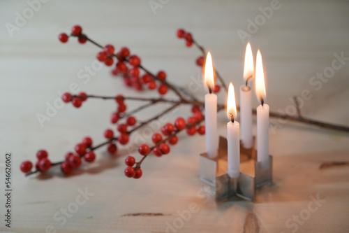 Mini advent wreath, four small candles on a cookie cutter in star shape in front of a holly branch with red berries on a wooden table, selected focus