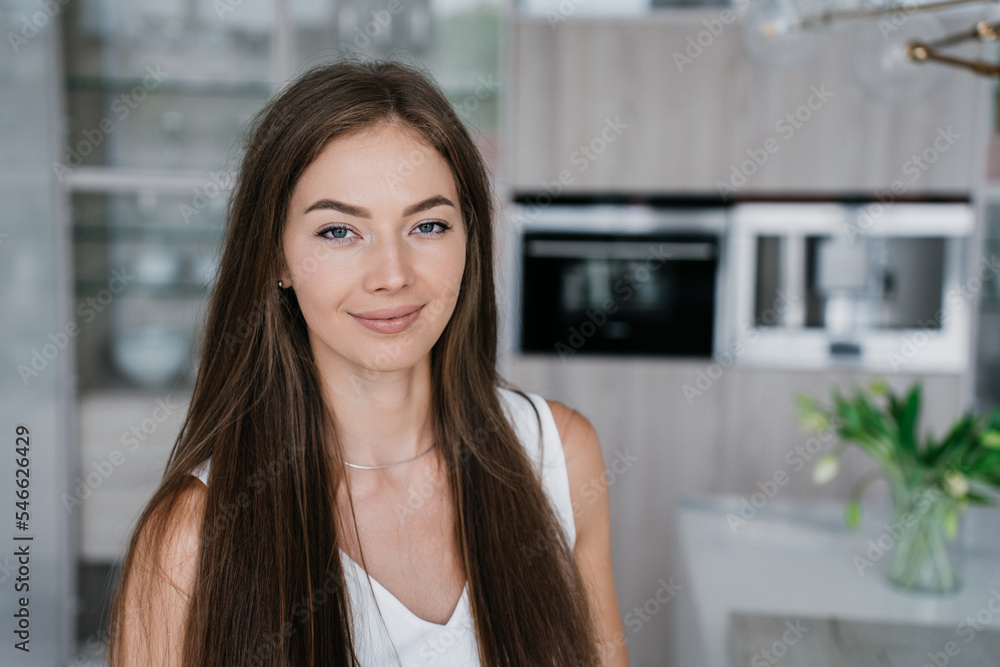 Close up portrait of confident blonde Caucasian girl with loose long hair looking at camera, smiles stands against blurry kitchen. Successful Italian beautiful businesswoman with perfect skin at home.