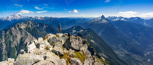 Adventurous athletic female hiker standing on top of a rugged mountain in the Pacific Northwest with jagged mountains in the background. 