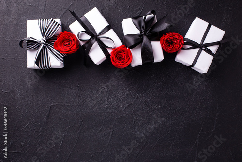  Romantic postcard. Border from wrapped boxes with presents and bright red roses flowers on black textured background. Place for text. Top view. Selective focus is on boxes.