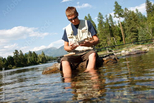 A teenage boy sits on a rock as he chooses a fly while fly fishing on the Swan River near Bigfork, Montana. photo