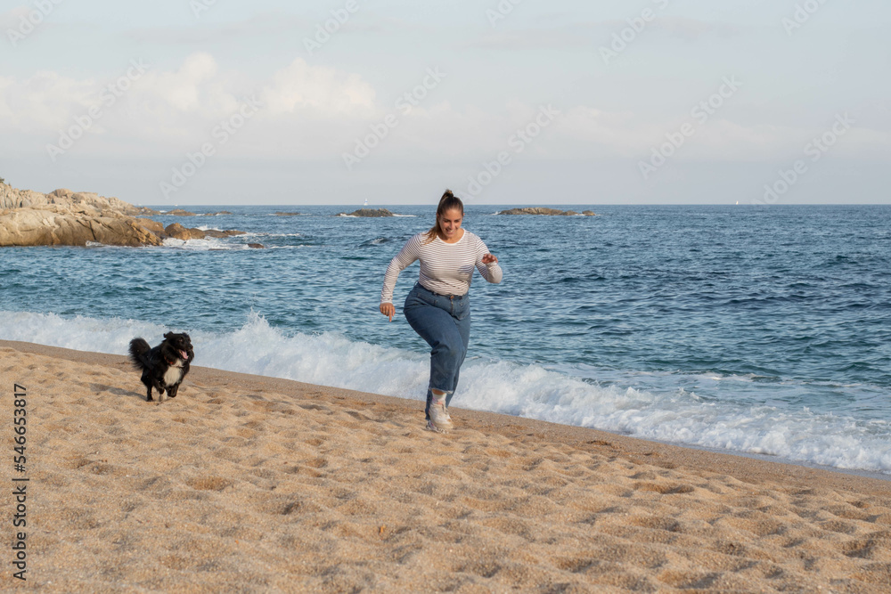 Mujer joven jugando en la playa con su bonito perro negro llamado Loki. Corriendo, sonriendo, queriéndose y abrazándose en actitud feliz y disfrutando de un agradable día - obrazy, fototapety, plakaty 