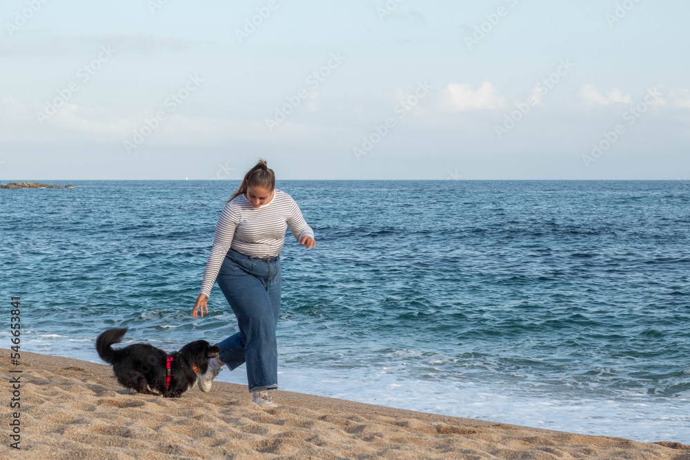 Mujer joven jugando en la playa con su bonito perro negro llamado Loki. Corriendo, sonriendo, queriéndose y abrazándose en actitud feliz y disfrutando de un agradable día - obrazy, fototapety, plakaty 