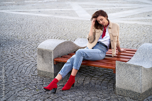 Beautiful young woman wearing teddy jacket jeans and white shirt sitting on a bench in the city