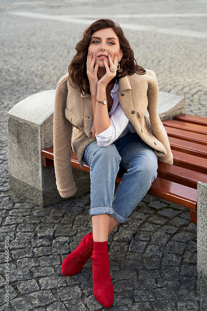 Beautiful young woman wearing teddy jacket jeans and white shirt sitting on a bench in the city