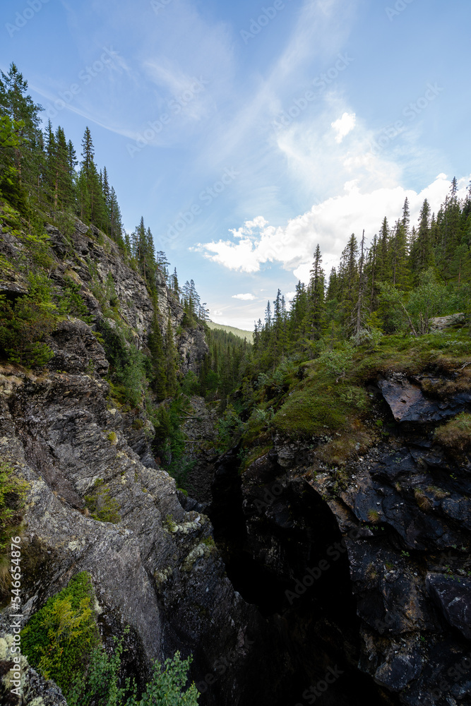 a blue dark narrow valley in Norway Helvete which has a forest with green trees along the edges, but above them opens a number of blue skies with puffy clouds a blue dark narrow valley in Norway Helvete which has a forest with green trees along the edges, but above them opens a number of blue skies with puffy clouds