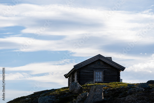 on gray stones overgrown with grass stands a small house with a checkered window, above the house there is a blue sky with floating light clouds