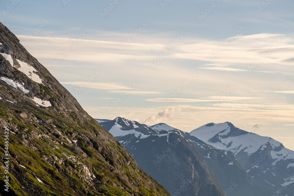 mountain landscape with a big deep valley that is between big mountains whose gray stone surface is covered with green moss, but on the top of the mountain there is snow and there is a beautiful sky