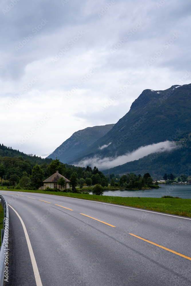 Naklejka premium a large gray asphalt road with a white line and a yellow solid line with a house with a fjord in the background and a mountain with a forest with white fog above it with clouds