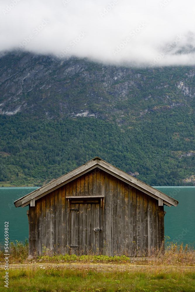 a gray small boat garage stands on the shore on a green meadow behind which is blue water above which a clan with a forest a gray small boat garage stands on the shore on a green meadow behind which is blue water above which a clan with a forest
