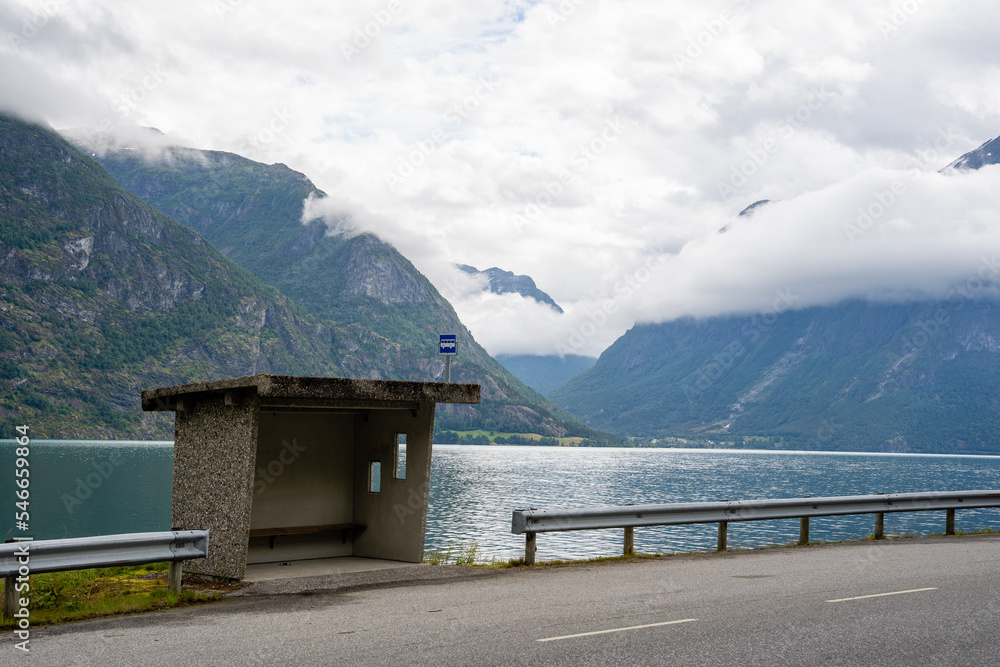 gray concrete bus stop on the side of the road behind which is a fjord with blue water and behind the fjord a beautiful view of big mountains covered with white fluffy clouds