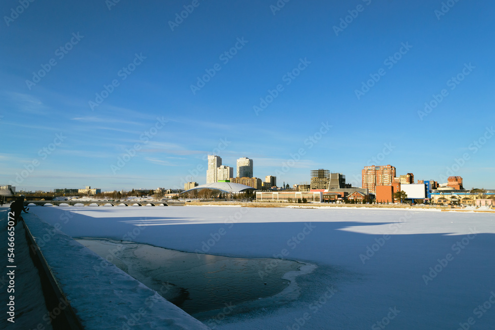 Panorama of a frozen river and buildings illuminated by the sun on a winter day against a blue sky