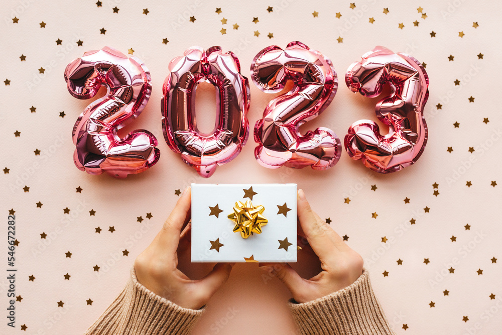 Fototapeta premium A person is holding a white gift box with a golden boy and a gift on a pink background. Next to confetti and gold and silver numbers 2023. Festive background of the concept of New Year and Christmas.