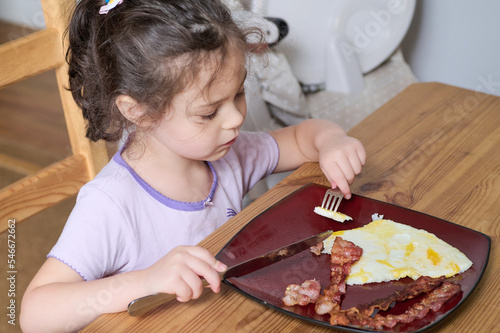 Young pretty girl eating eggs and bacon with knife and fork for the first time