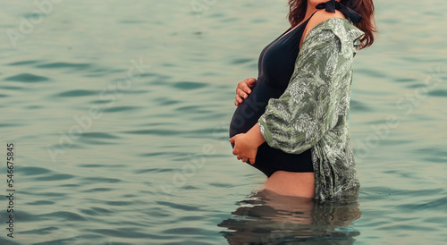 Fototapeta Naklejka Na Ścianę i Meble -  Motherhood and pregnancy. Young pregnant woman in swimsuit is standing in sea and holding her tummy. Copy space. Concept of water births