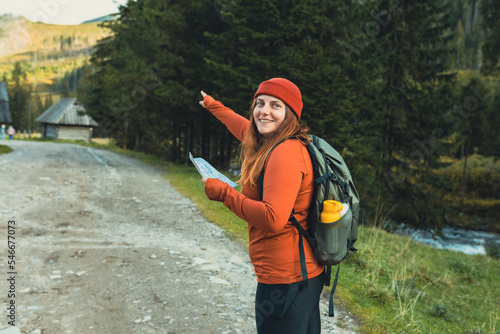 Stylish woman holding paper map relaxing in nature. Travel and active lifestyle concept. Hiking female lifestyle wearing backpack and red knit hat exercising outdoors. Girl pointing something
