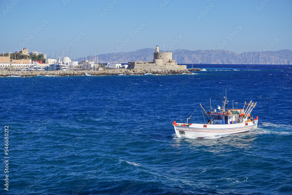 Rhodes, Greece: A fishing boat passes St. Nicholas Lighthouse (1863) in St. Nicholas Fortress in Mandraki Harbor on the Aegean island of Rhodes.