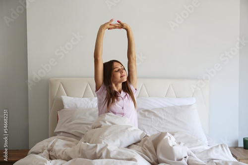 Young beautiful woman stretching in her bed after waking up in the morning. Sleepy brunette female with satisfied facial expression. Healthy sleeping habits concept. Close up, copy space, background. photo