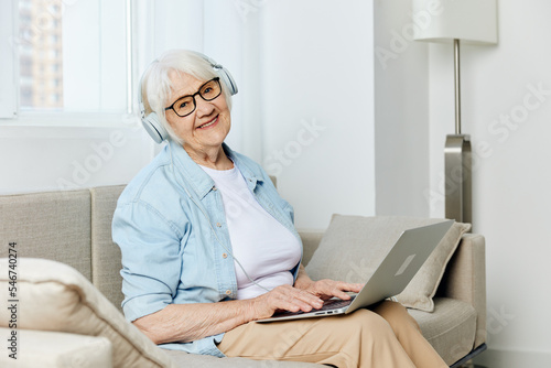 a happy elderly lady is sitting on the sofa in a light shirt and holding a laptop on her lap, smiling happily while looking at the camera with glasses on her face and headphones on her head