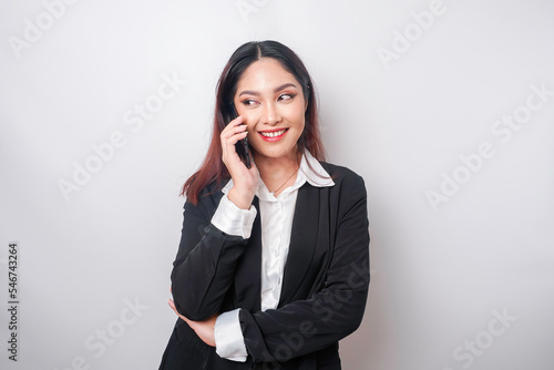 A portrait of a happy Asian businesswoman is smiling while talking on phone call wearing a black suit isolated by a white background
