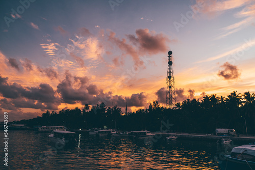Wide-angle shot of a stunning sunset sky over the an island jetty full of small passenger boats, motorboats, and launches, with silhouette of palm trees and a communications tower in the background