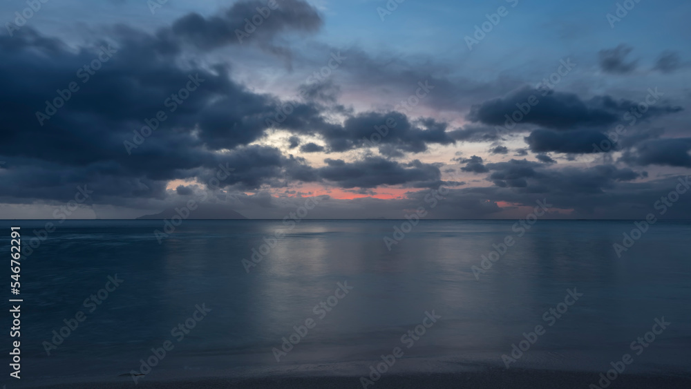 The evening sky over the calm turquoise ocean is highlighted in pink. Blue clouds. Long exposure. Seychelles