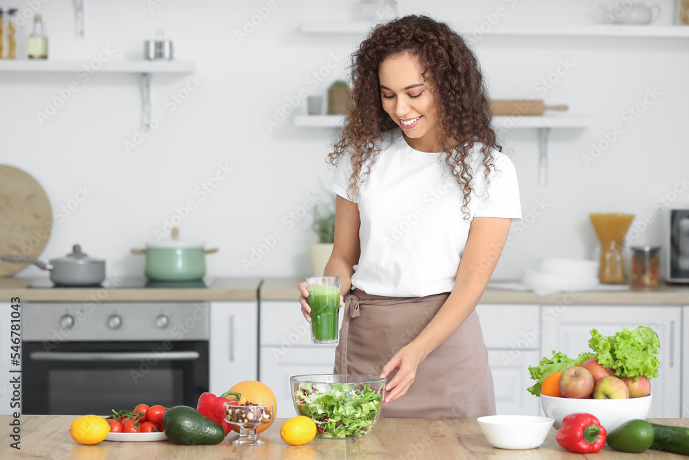 Young African-American woman with healthy salad and smoothie in kitchen