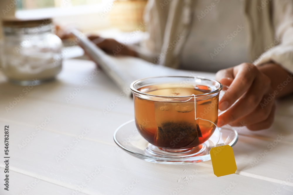 Woman holding glass cup with tea bag at white wooden table, closeup. Space for text Woman holding glass cup with tea bag at white wooden table, closeup. Space for text
