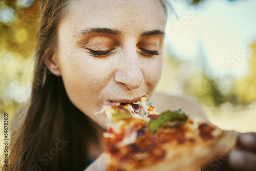 Eating  pizza and hungry with a woman biting a slice of fast food outdoor in a park or garden on the weekend. Food  bite and lunch with a young female enjoying a snack outside for her hunger