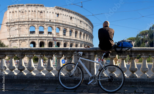 Photography man sitting on low wall looks at the sea with bike in Rome with Colosseo