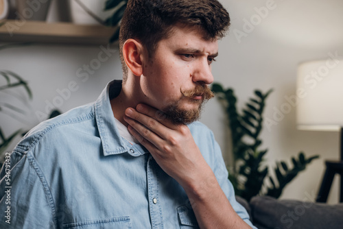 Sick young caucasian man sitting on couch at home touch neck suffer from angina has sore throat unhealthy guy painful swallow feel discomfort coronavirus disease symptom respiratory infection
