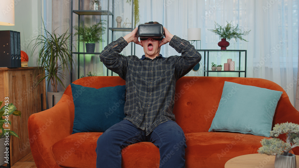 Young man use VR headset glasses at home, enjoying 3D 360 video concept, entertaining, in positive mood, moving hands in air, sitting on sofa in living room. Virtual reality, modern gadget device
