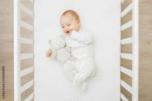 Adorable happy baby boy in white bodysuit hugging teddy bear and lying down on back on mattress in crib at home room. 6 months old infant playing with first friend. Closeup. Top down view.