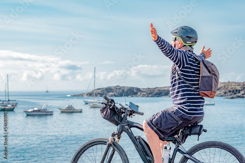 Happy cyclist man runs with electric bike next to the sea looking at the horizon with open arms , smiling senior male enjoys freedom and healthy lifestyle during retirement