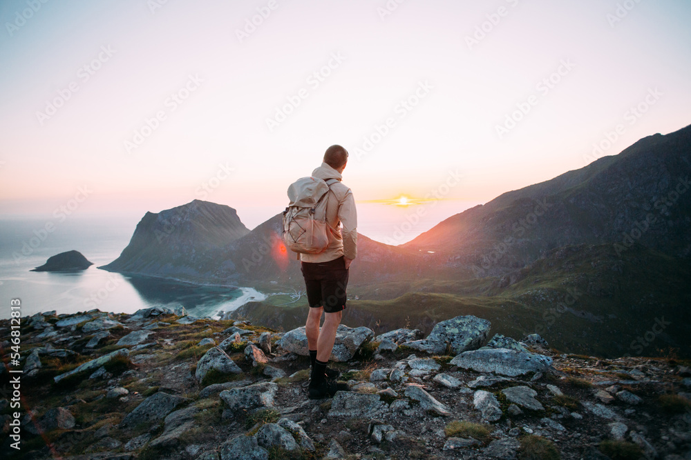 Obraz premium Hiker stand on top of mountain climb. Man in hiking outfit with backpack stand on top of hill overlooking epic mountain views covered by clouds. Hiking in Norway, Lofoten