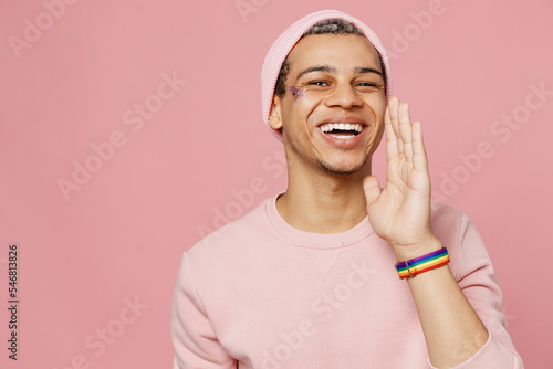 Young promoter gay man wears sweatshirt hat scream hot news about sales discount hold hand near mouth isolated on plain pastel light pink color background studio portrait Lifestyle lgbtq pride concept photo