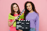 Young two friends smiling cheerful fun women 20s wears green purple shirts together holding classic black film making clapperboard isolated on pastel plain light pink color background studio portrait.